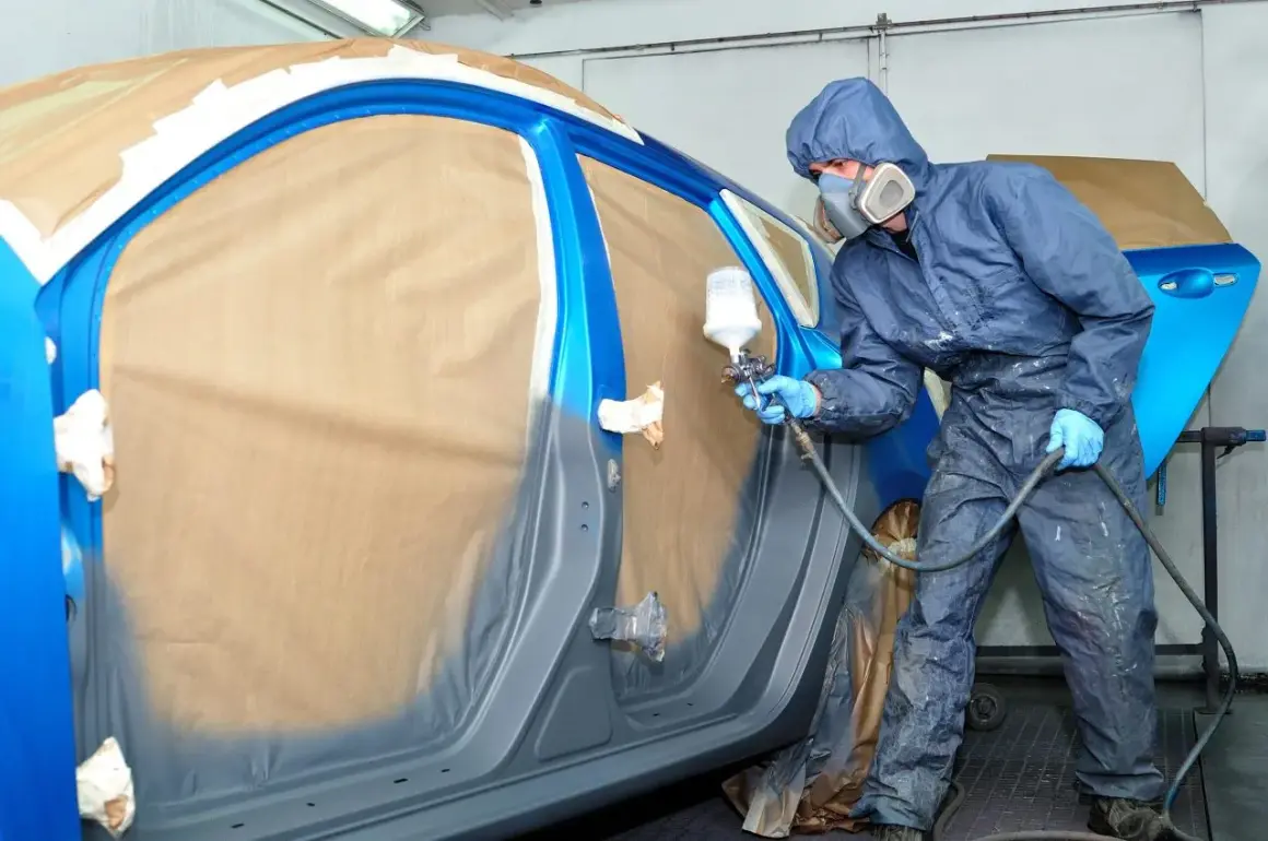 Pittore in tuta blu spruzza vernice su un'auto blu. Il costo per cambiare il colore della macchina sul libretto è un dettaglio importante.