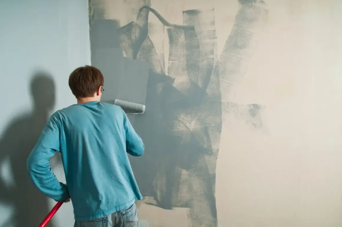Un uomo con una maglietta blu sta dipingendo un muro con un rullo. La pittura si stacca dal muro in alcune zone, creando un effetto astratto.