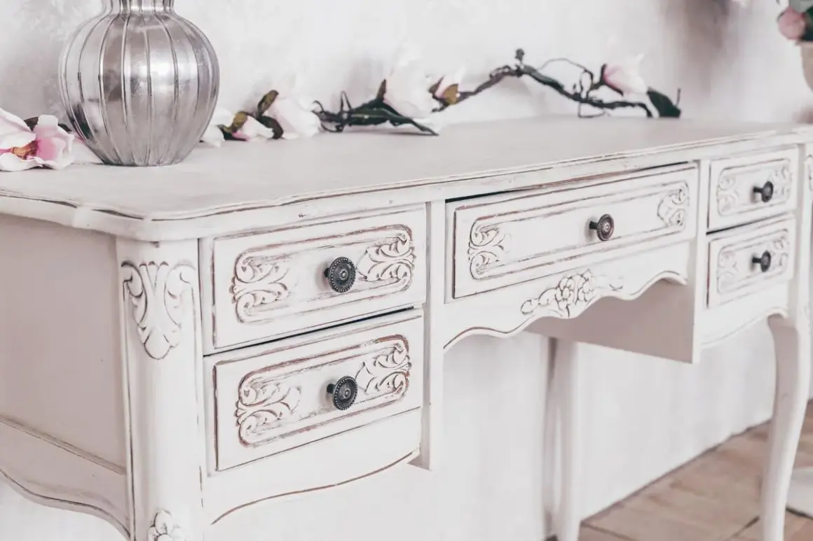 Un mobile da marrone a bianco, con dettagli intagliati e pomelli antichi. Un vaso d'argento e fiori decorano il piano.