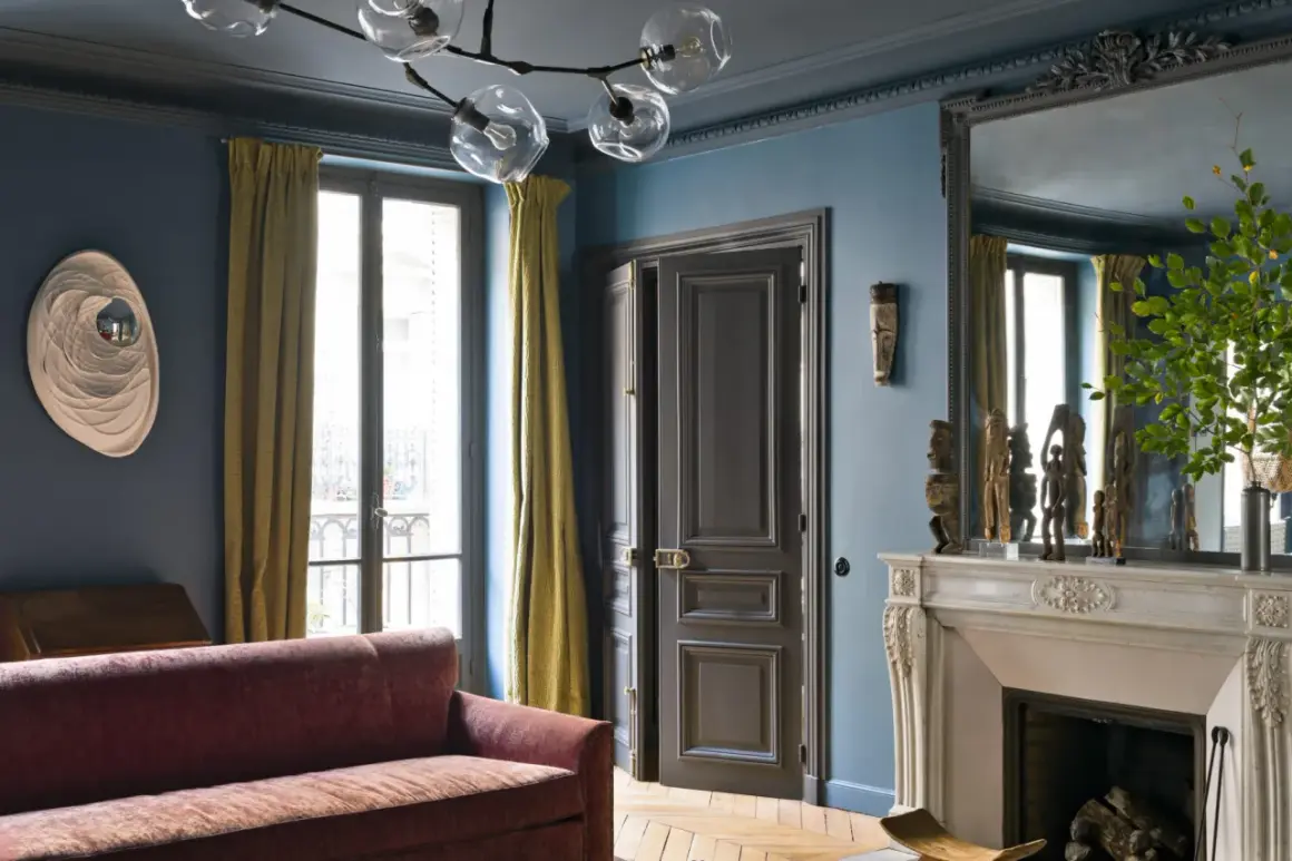 Soggiorno elegante con pareti blu balena Pantone, divano bordeaux, tendaggi ocra e camino in marmo.