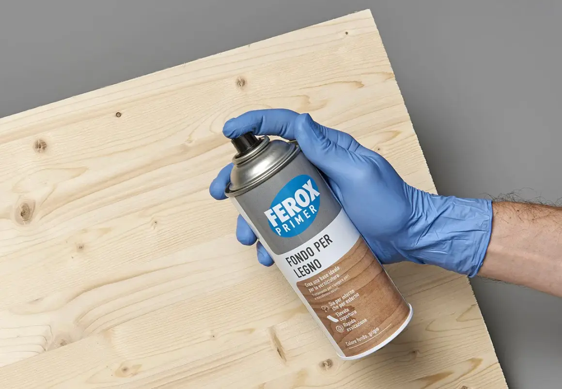 Mano con guanto blu tiene bomboletta spray "Ferox Primer Fondo per Legno", pronta per preparare la superficie.