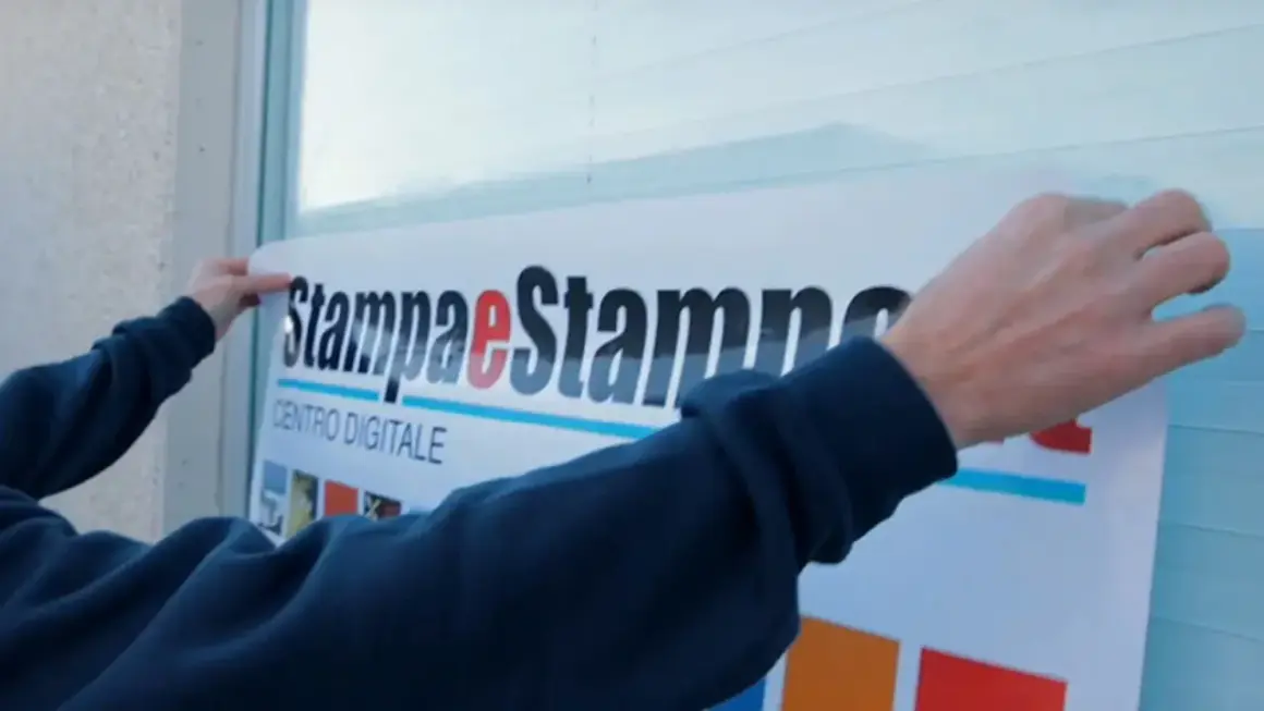 Mani applicano un grande adesivo con logo "Stampa e Stampe Centro Digitale" su una superficie, mostrando come applicare un adesivo senza bolle per un risultato perfetto.