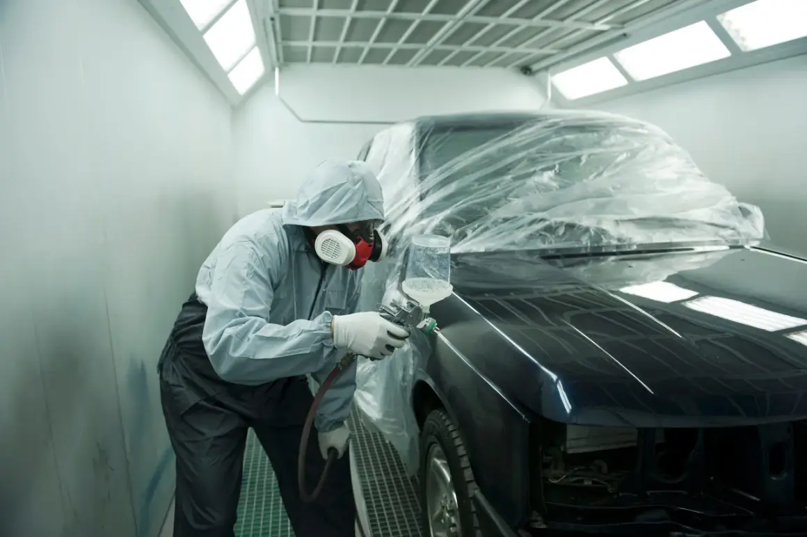 Un operaio in tuta protettiva e maschera spruzza vernice su un'auto scura, parzialmente coperta da un telo trasparente. L'auto sembra rovinata, in fase di restauro.