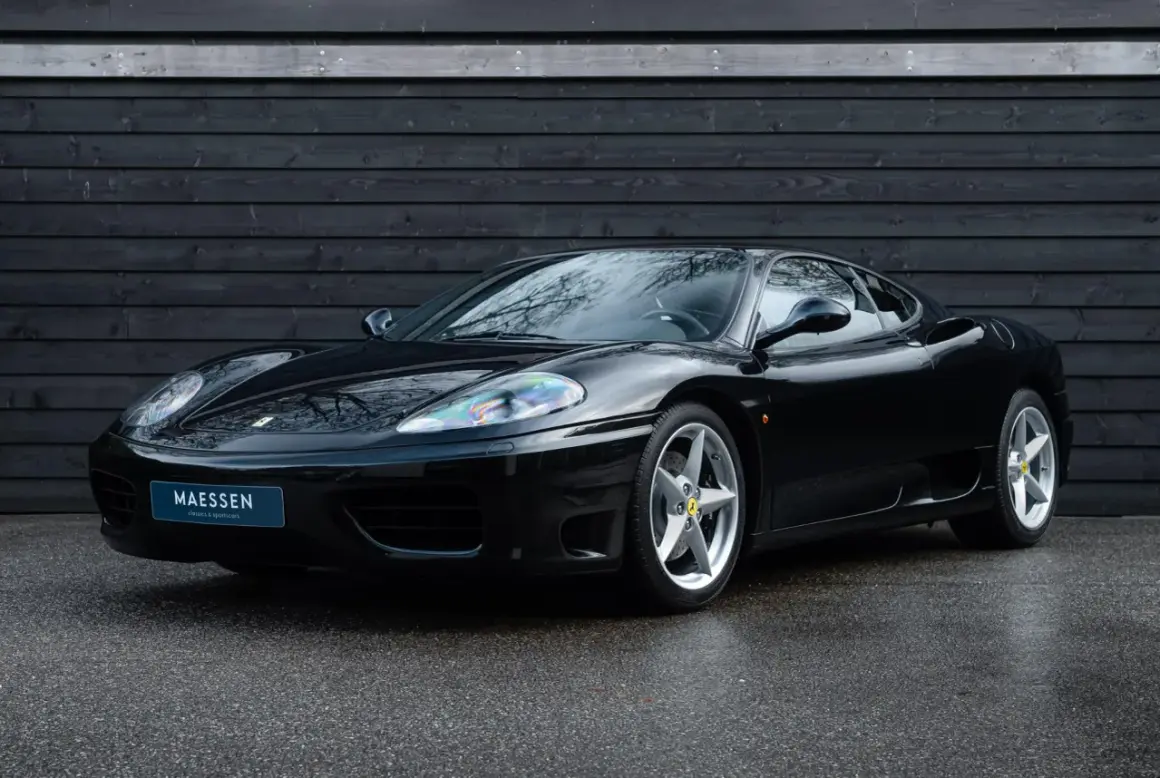 Una splendida auto nera, una Ferrari 360 Modena, parcheggiata su asfalto bagnato davanti a un muro di legno scuro.