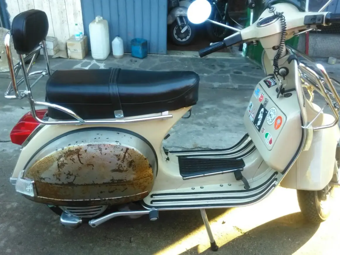 Vespa color crema con evidente effetto ruggine sul parafango posteriore. La sella è nera, con portapacchi cromato.