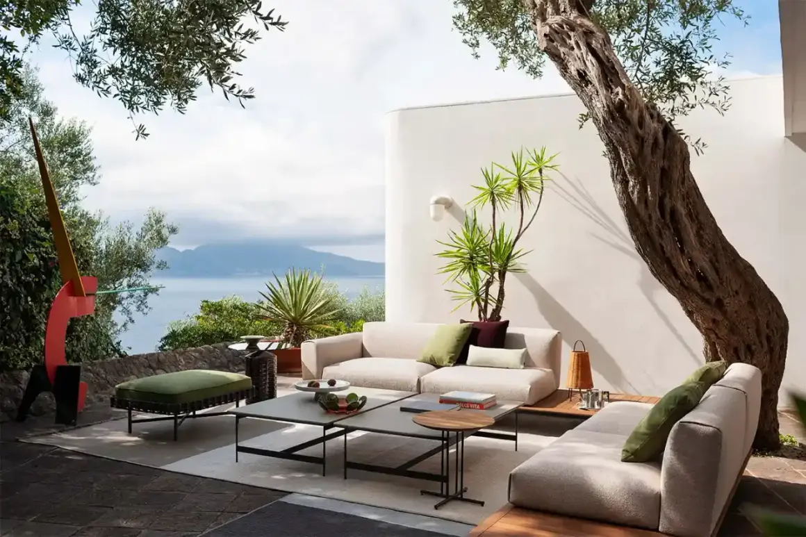 Arredare terrazzo con divani chiari, tavolini grigi e un pouf verde, con vista mare e scultura colorata.