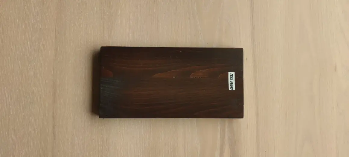 Tavola scura con evidenti venature del legno, su sfondo chiaro con venature simili. Etichetta bianca con scritta "NOCE MEDIO".