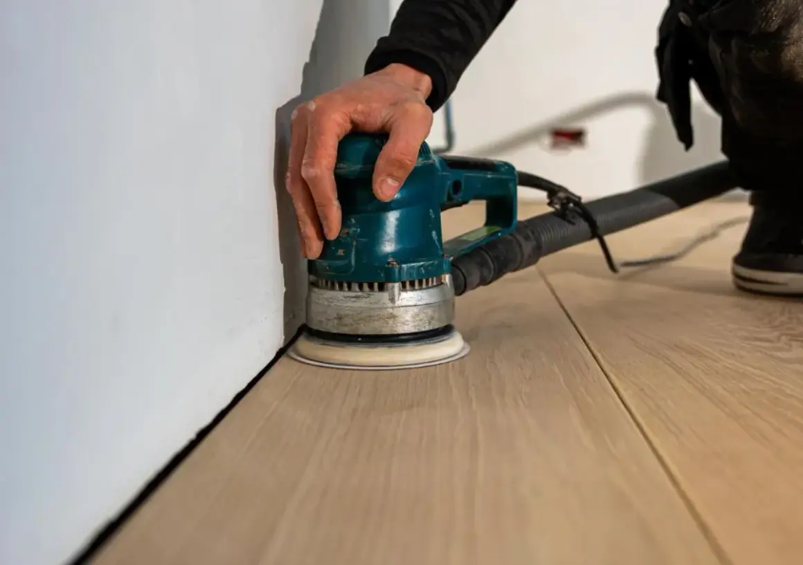 Mano che guida una levigatrice orbitale per la lamatura meccanica di un nuovo pavimento in legno, vicino a un muro bianco.