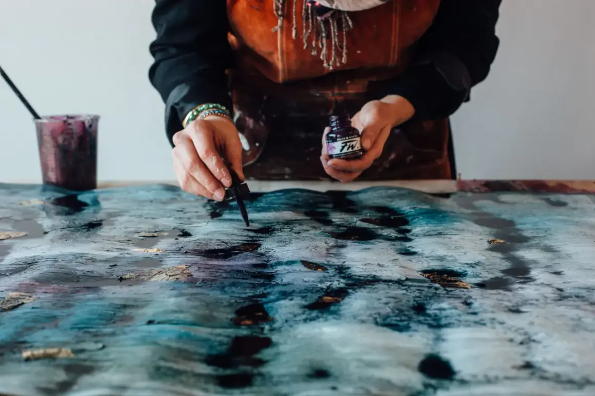 Mani che creano un'opera d'arte con colori acrilici su plastica, gocciolando inchiostro viola su una superficie texturizzata blu e oro.