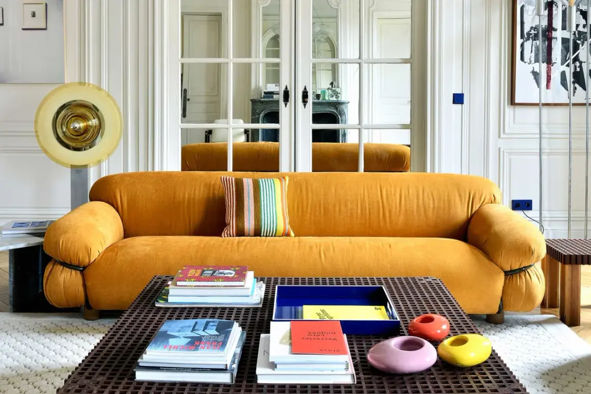 Divano senape con cuscino a righe colorate. Tavolino con libri e oggetti decorativi. Abbinamenti moderni e audaci.