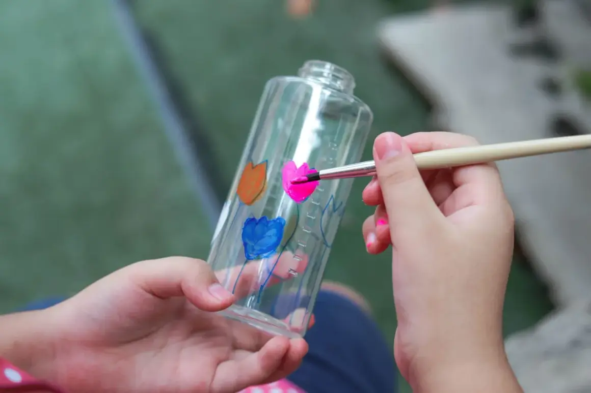 Bambino dipinge fiori su una bottiglia trasparente, un modo creativo per imparare come colorare la plastica trasparente.