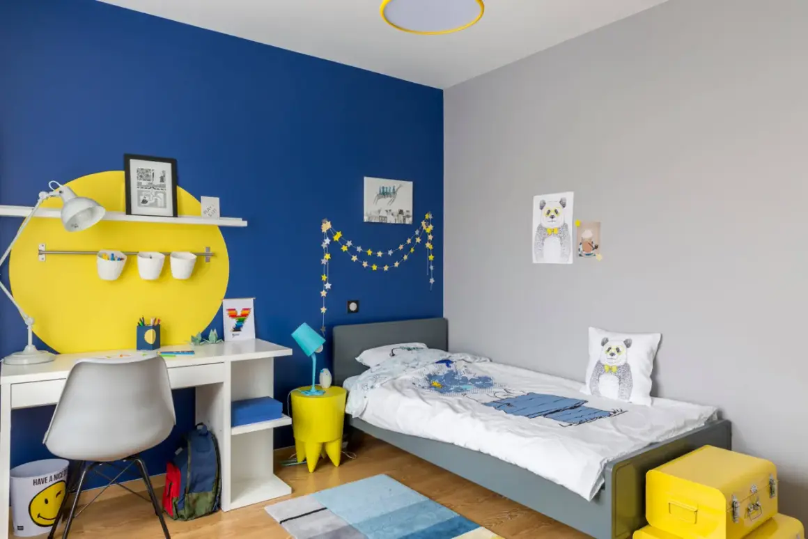 Cameretta con parete blu vivace e decorazioni a stelle. Scrivania bianca, letto grigio, cuscino panda. Ottime idee pittura cameretta per un ambiente allegro.