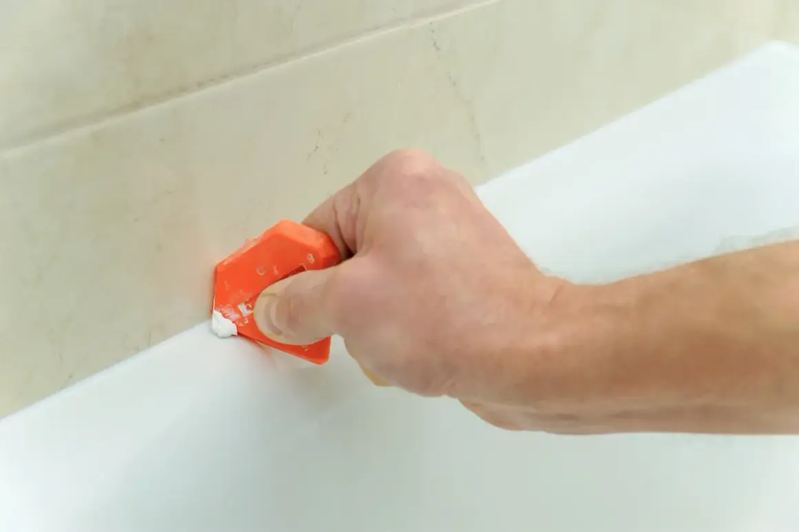 Mano usa spatola arancione per rifinire silicone, un lavoro antisilicone fai da te per sigillare la vasca.