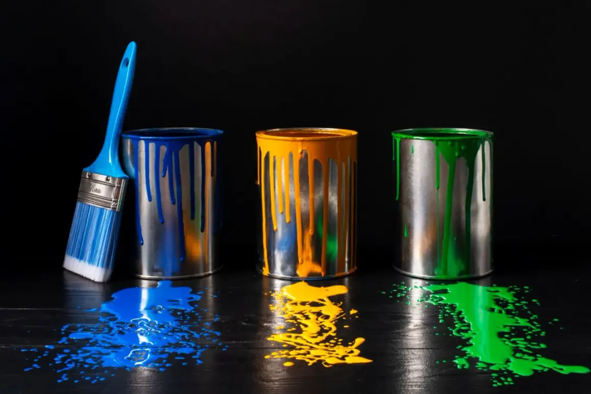Tre barattoli di vernice (blu, giallo, verde) con gocce e schizzi su un tavolo scuro. Un pennello blu accanto. Il solvente cos'è?