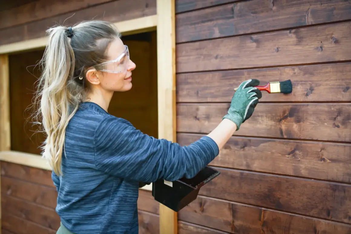 Donna con occhiali protettivi applica una vernice scura su una parete di legno, un tipo di pittura che protegge e abbellisce.