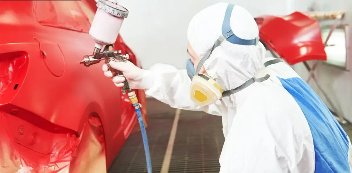 Persona in tuta protettiva e maschera effettua verniciare auto fai da te con pistola a spruzzo su un'auto rossa.