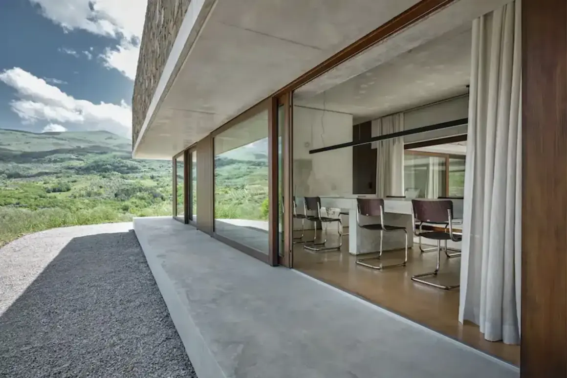 Interno moderno con tavolo e sedie, vista su paesaggio verde. Architettura in stile brutalista con cemento a vista.