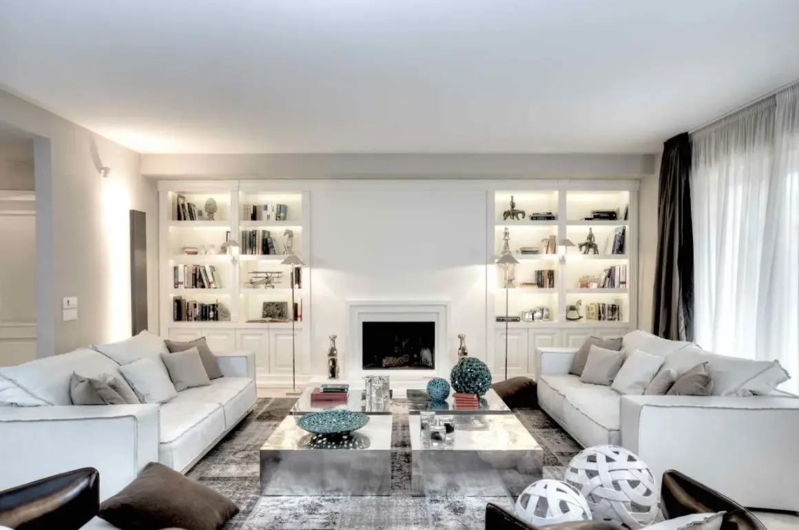 Soggiorno elegante con divani bianchi, librerie illuminate e camino. Le pareti color bianco latte creano un'atmosfera luminosa e ariosa.