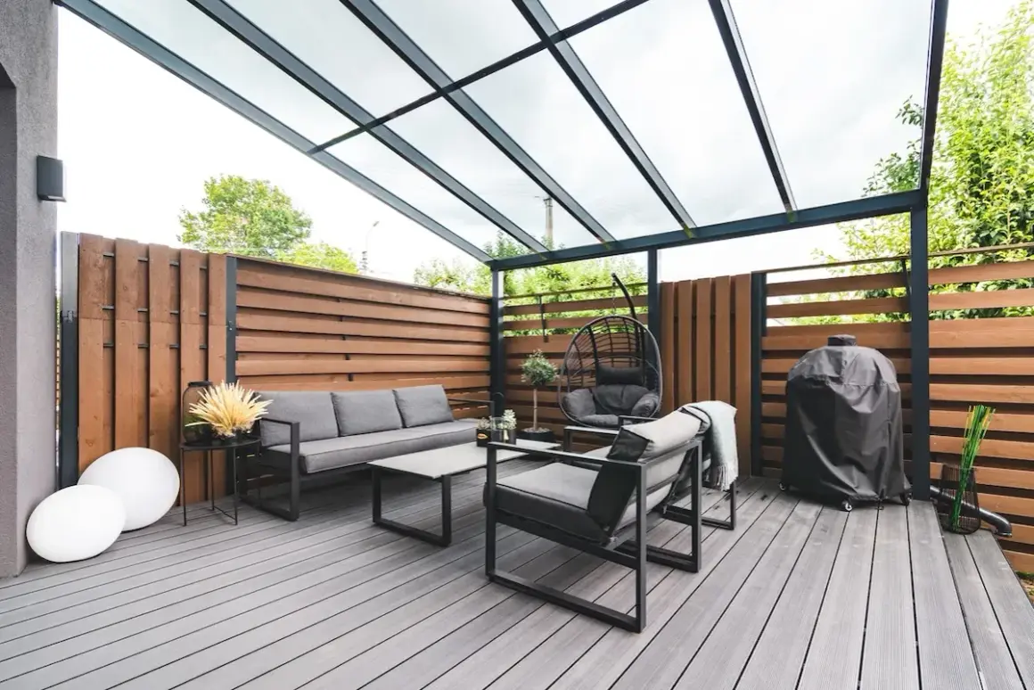 Arredamento moderno per un balcone con idee frangivista: divano, poltrone, dondolo e barbecue sotto una tettoia trasparente.