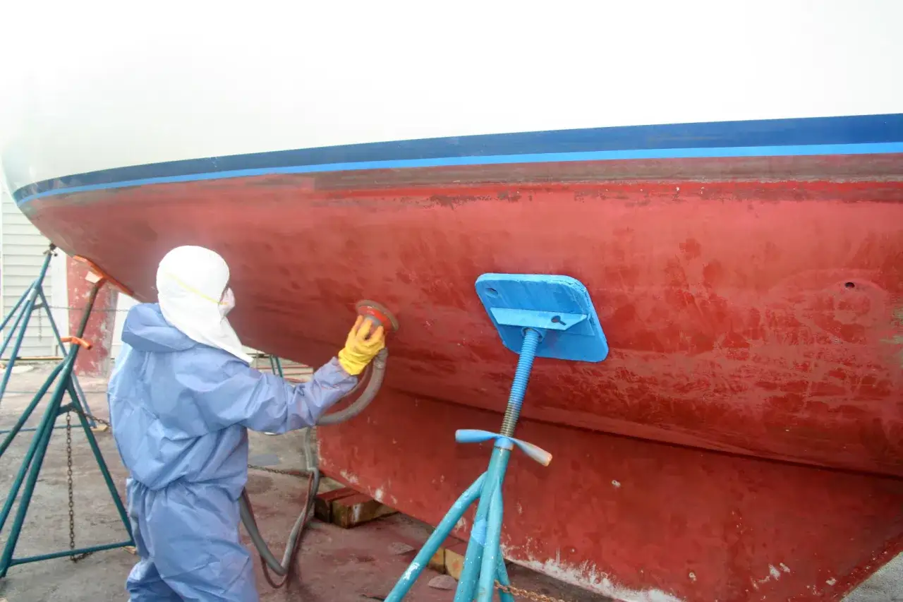 Persona in tuta protettiva leviga lo scafo di una barca, rimuovendo il vecchio antivegetativo per prepararlo a una nuova applicazione.