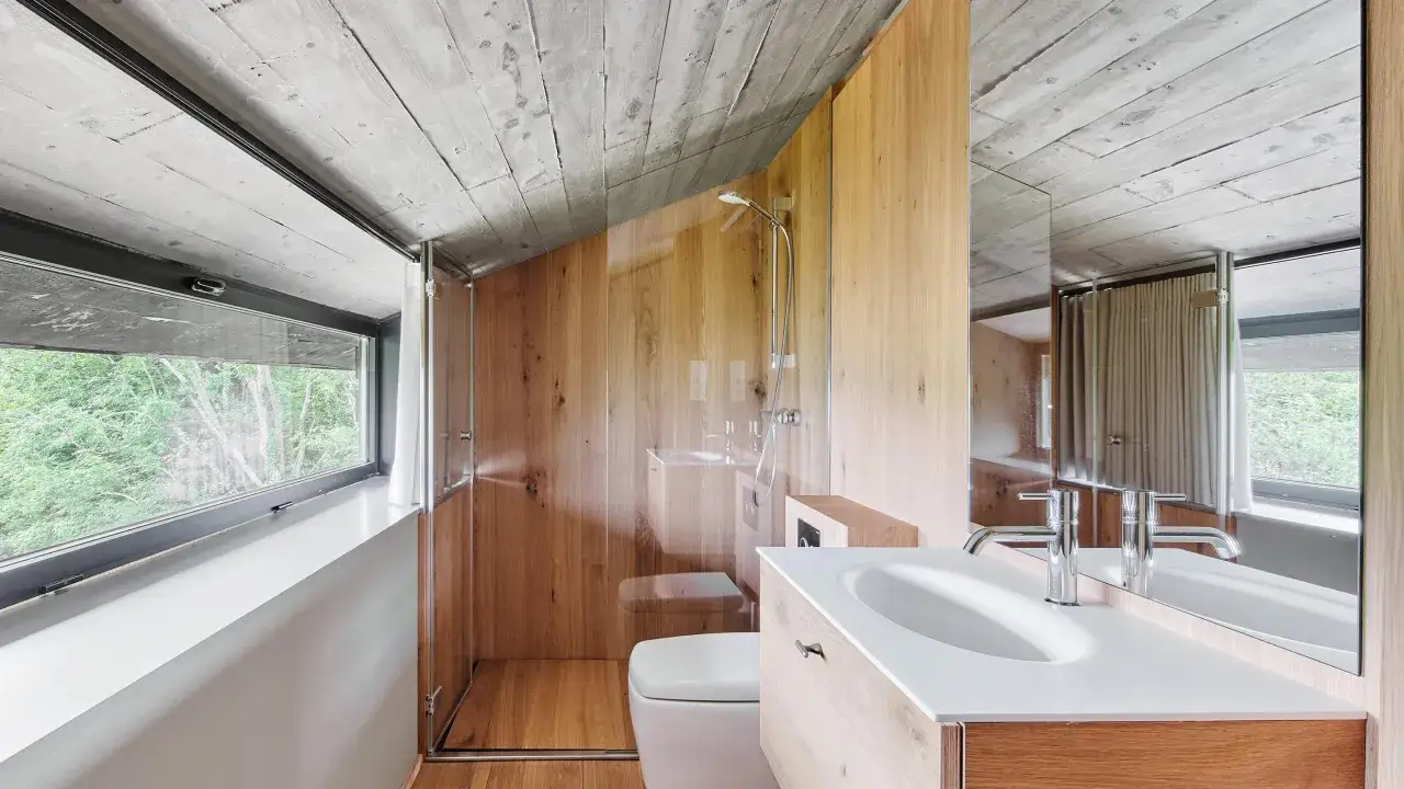Bagno moderno con box doccia in legno fai da te, lavabo e WC. Soffitto in cemento grezzo e ampia finestra con vista sul verde.