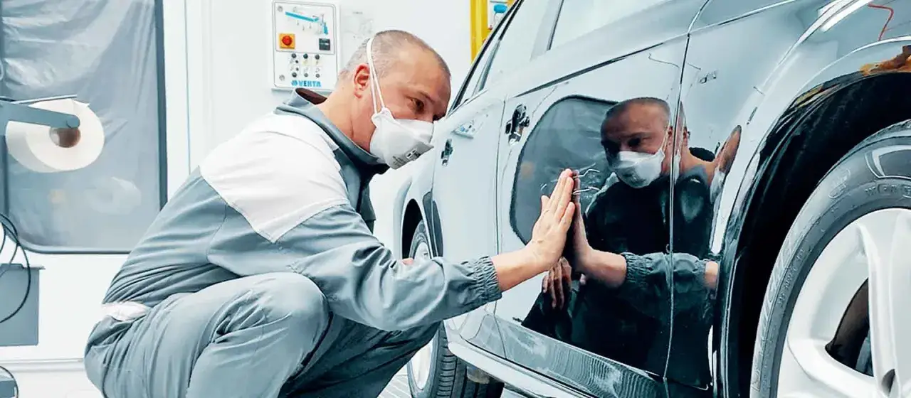 Un operaio con mascherina controlla la carrozzeria lucida di un'auto, pronto per carteggiare auto e rifinire la verniciatura.