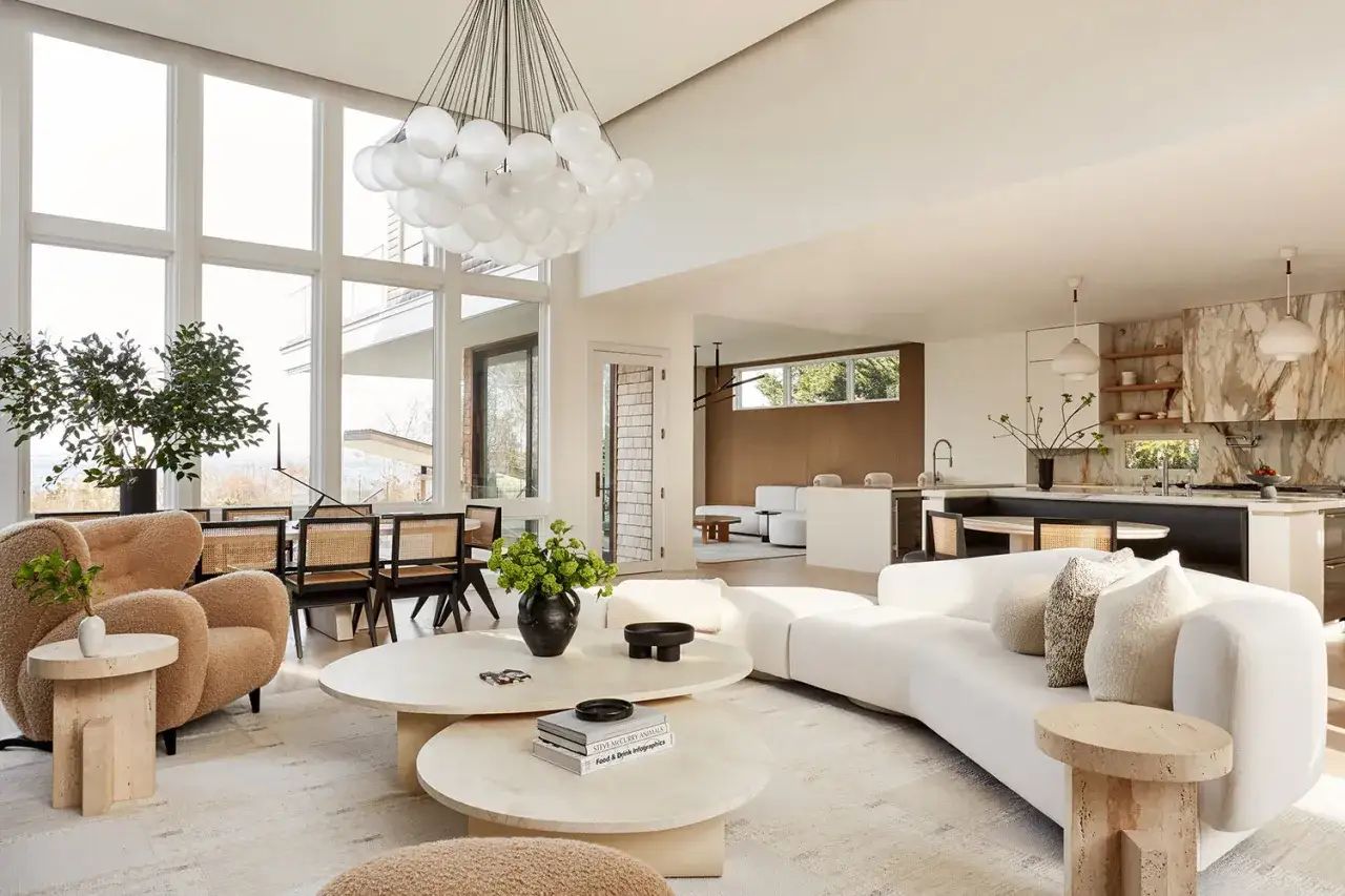 Soggiorno luminoso con divano curvo bianco, tavolini in travertino e poltrona in bouclé. L'arredamento è un perfetto esempio di stile organic modern, con materiali naturali e forme morbide.