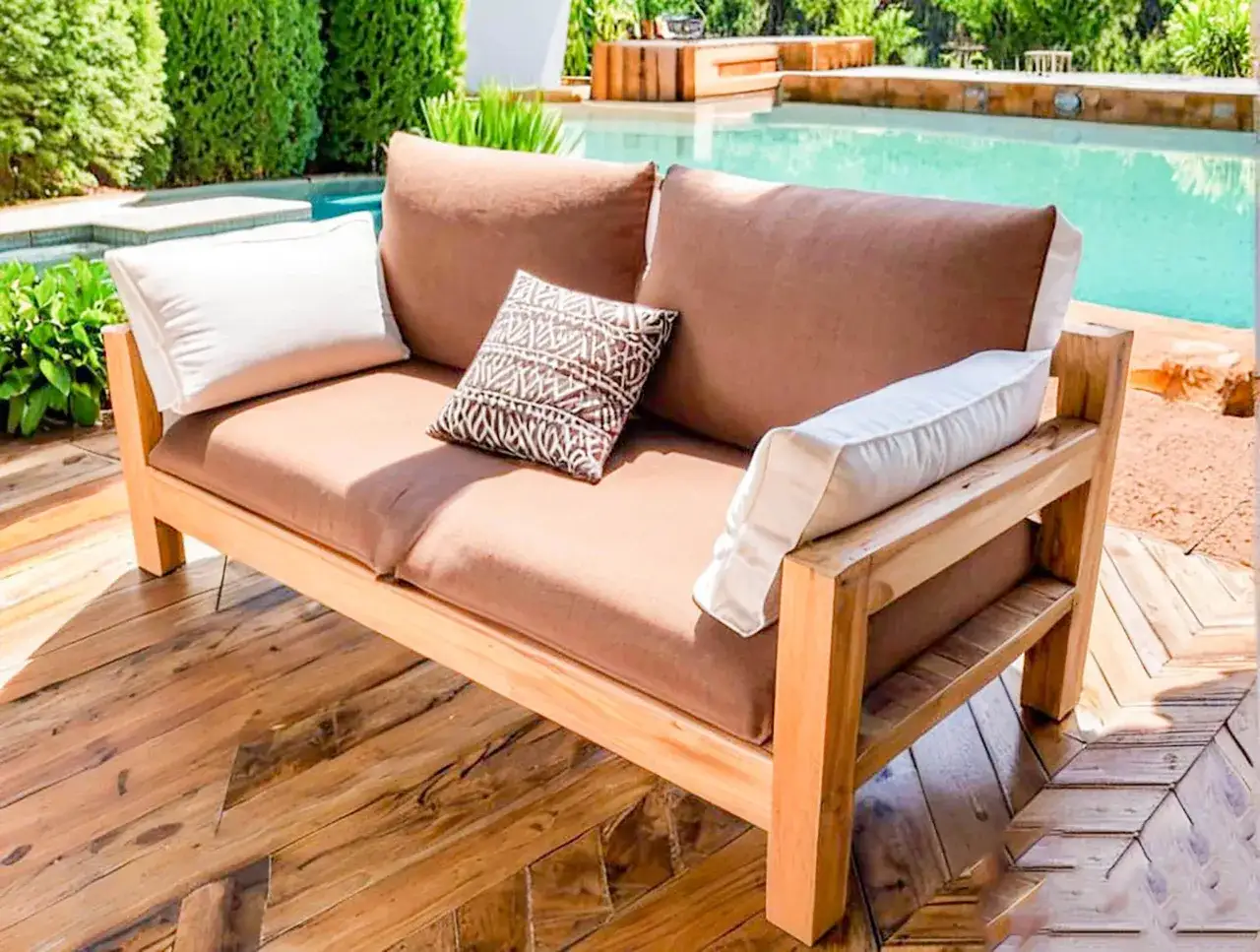Divano fai da te in legno con cuscini marroni e bianchi, posizionato su una terrazza in legno accanto a una piscina circondata da vegetazione.