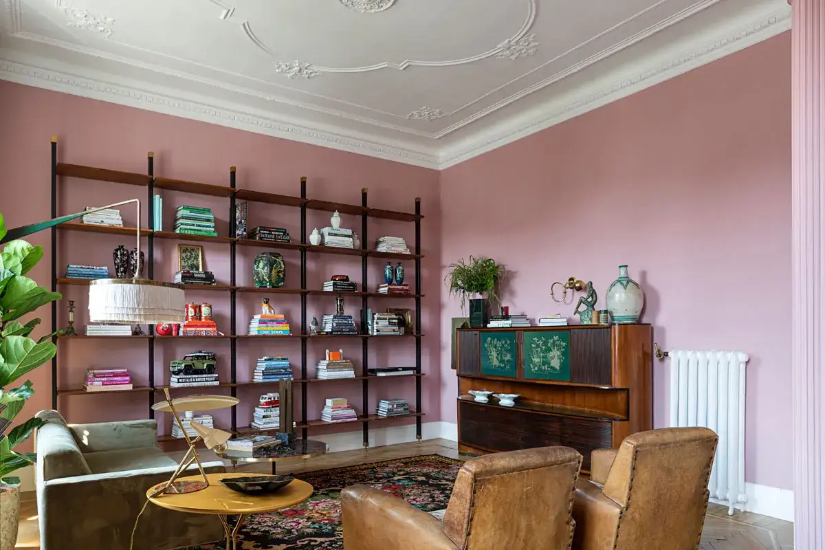 Salotto accogliente con pareti rosa cipria, libreria piena di libri e oggetti, mobili in legno scuro e poltrone in pelle. I colori caldi creano un'atmosfera rilassante.
