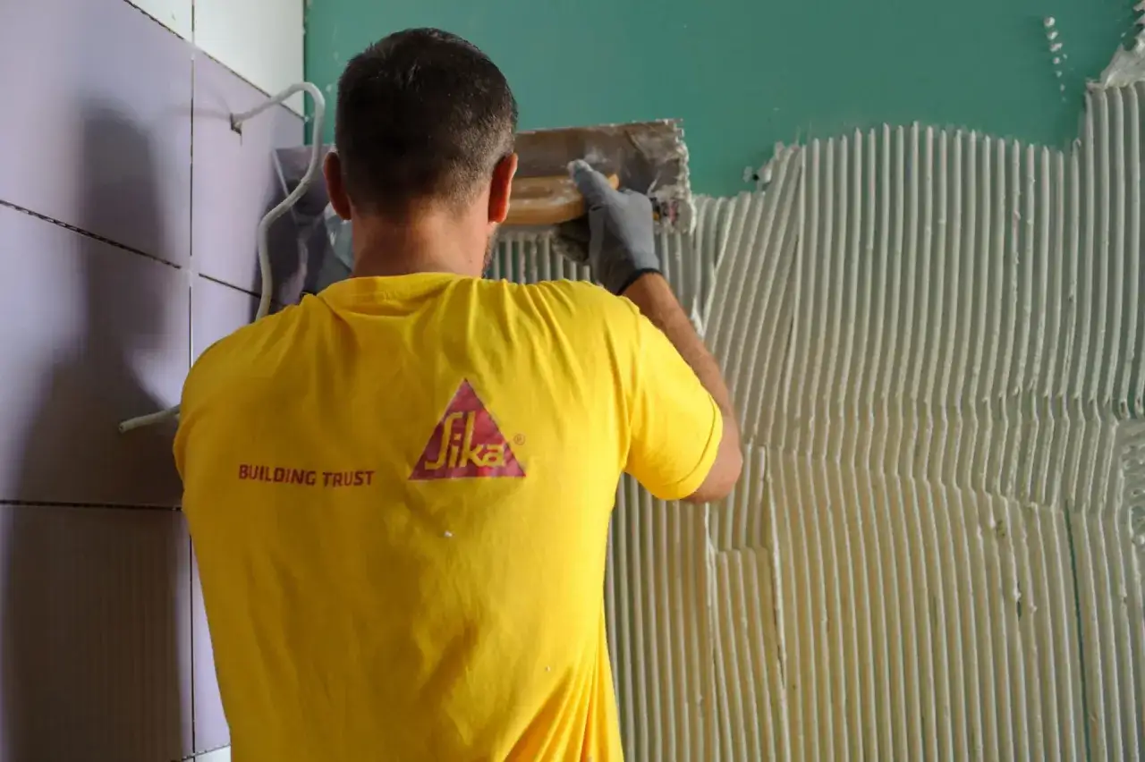 Operaio con maglietta gialla Sika applica colla sul muro, mostrando come attaccare piastrelle.