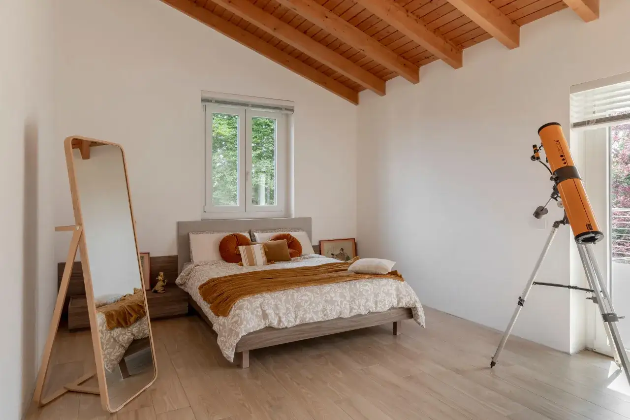Camera da letto con parquet chiaro, letto con copriletto bianco e arancione, specchio e telescopio.