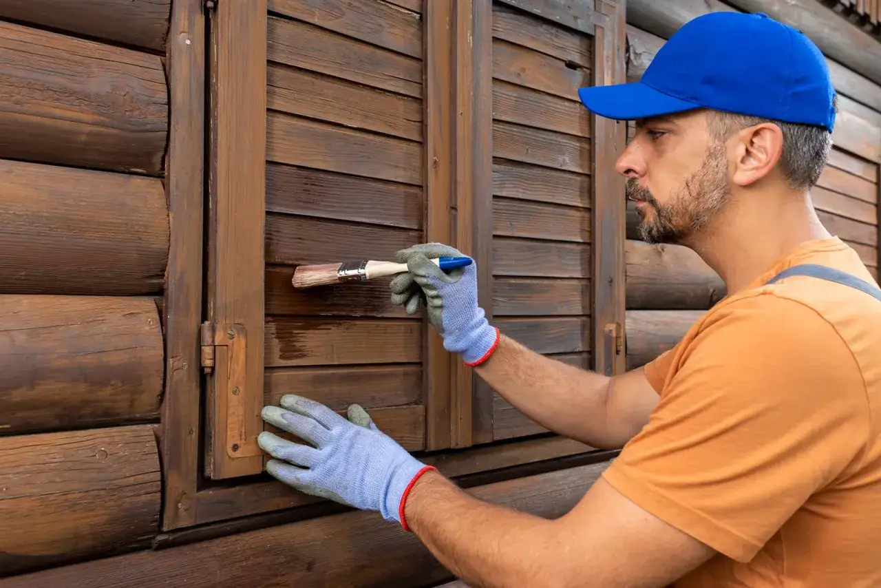 Uomo con cappellino blu e guanti azzurri effettua la verniciatura persiane in legno, rinnovando la facciata di una casa in tronchi.