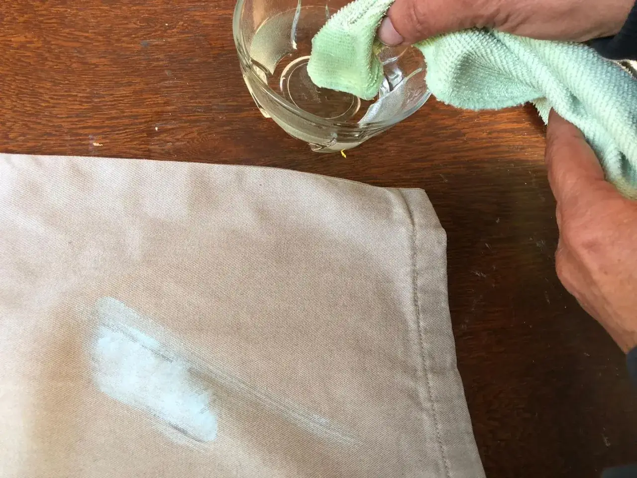 Come togliere macchie di vernice secca dai vestiti: un panno imbevuto in un bicchiere d'acqua tampona una macchia chiara su tessuto beige.