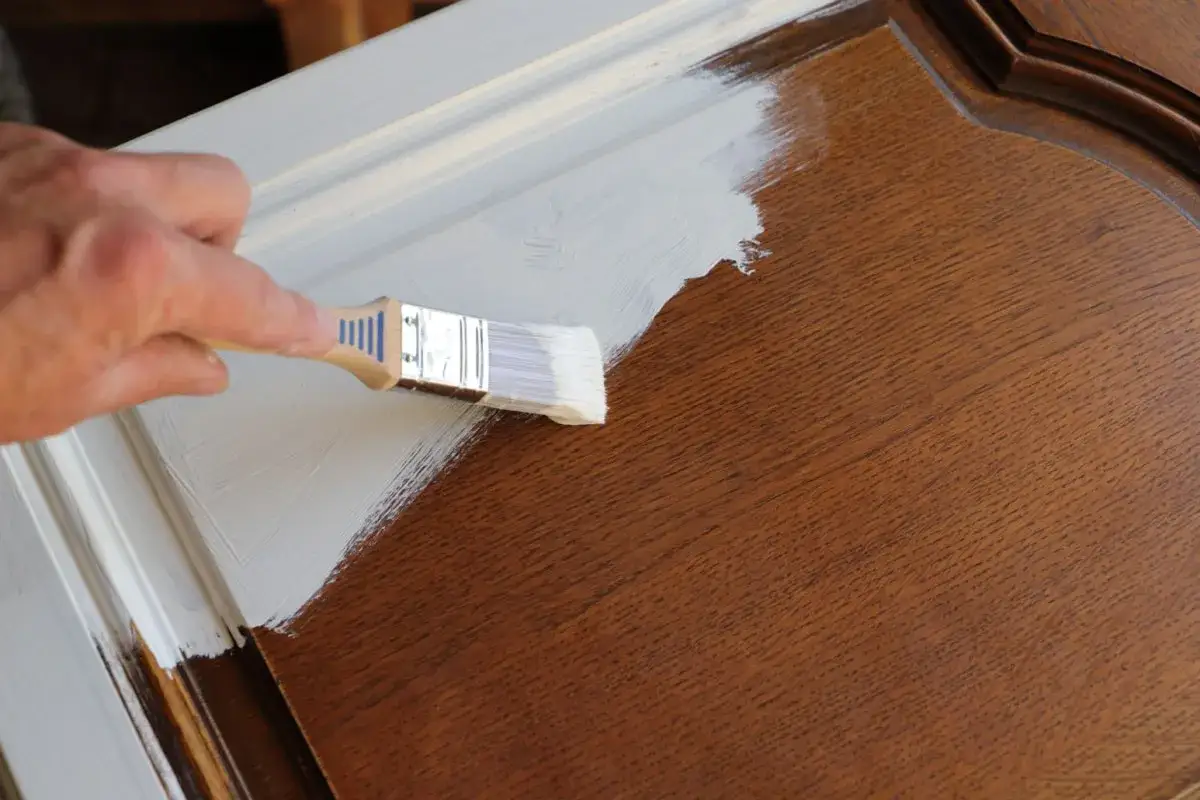 Mano che dipinge con pennello bianco su mobile in legno scuro. La cementite a cosa serve? A preparare la superficie per una nuova finitura.