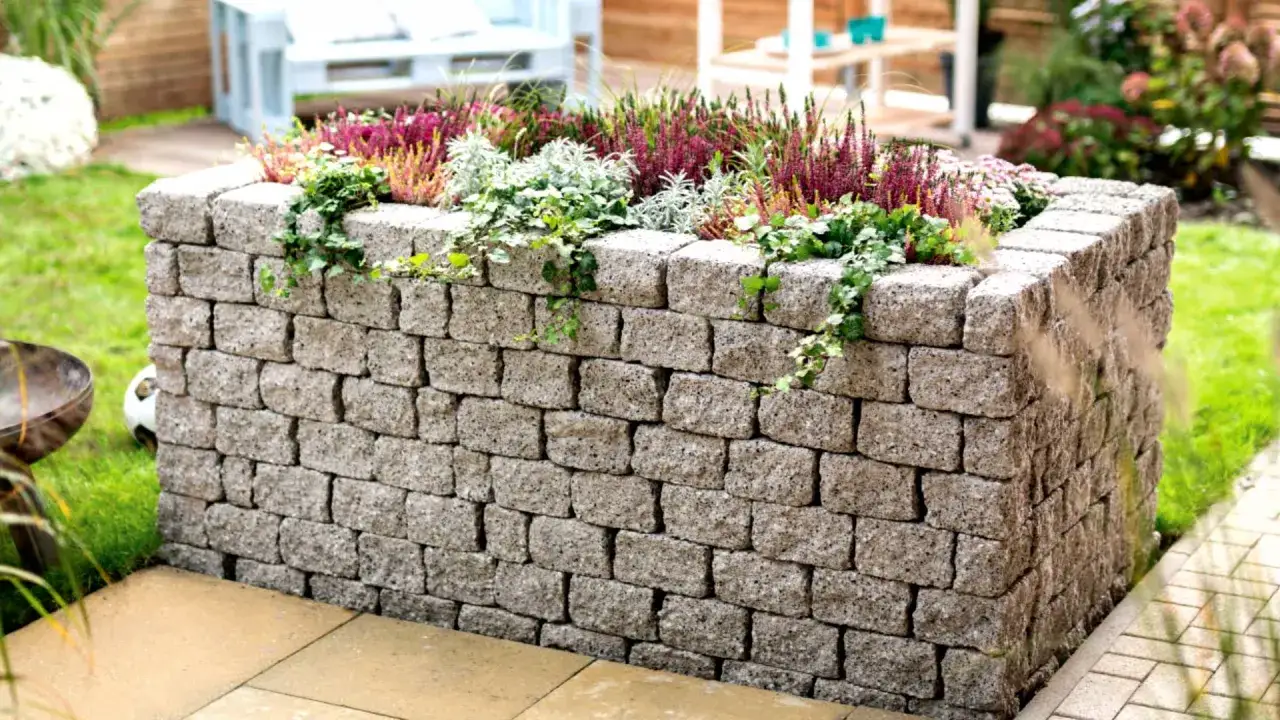Un rialzo in pietra per fiori, un'idea per muretto giardino che aggiunge un tocco rustico e moderno. Piante verdi e fiori viola decorano il bordo.