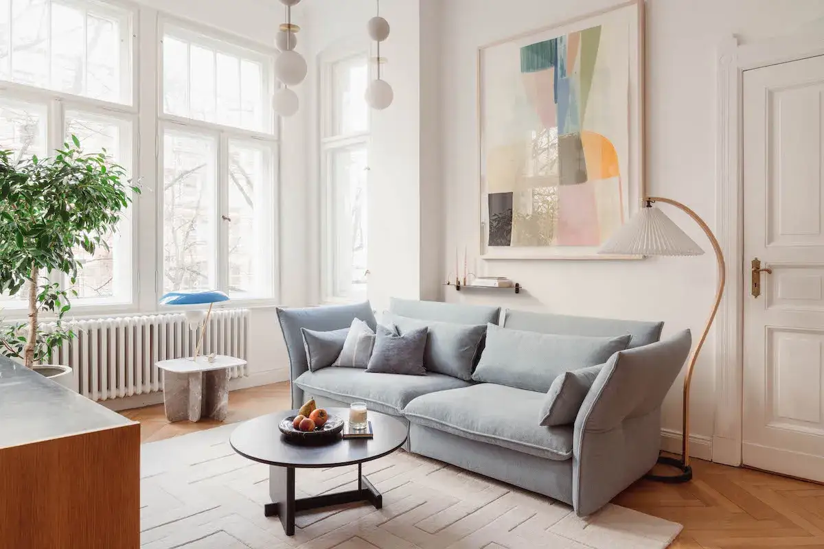 Soggiorno luminoso con divano azzurro, tavolino nero, quadro astratto e lampada ad arco. Il colore bianco codice domina le pareti e le ampie finestre.