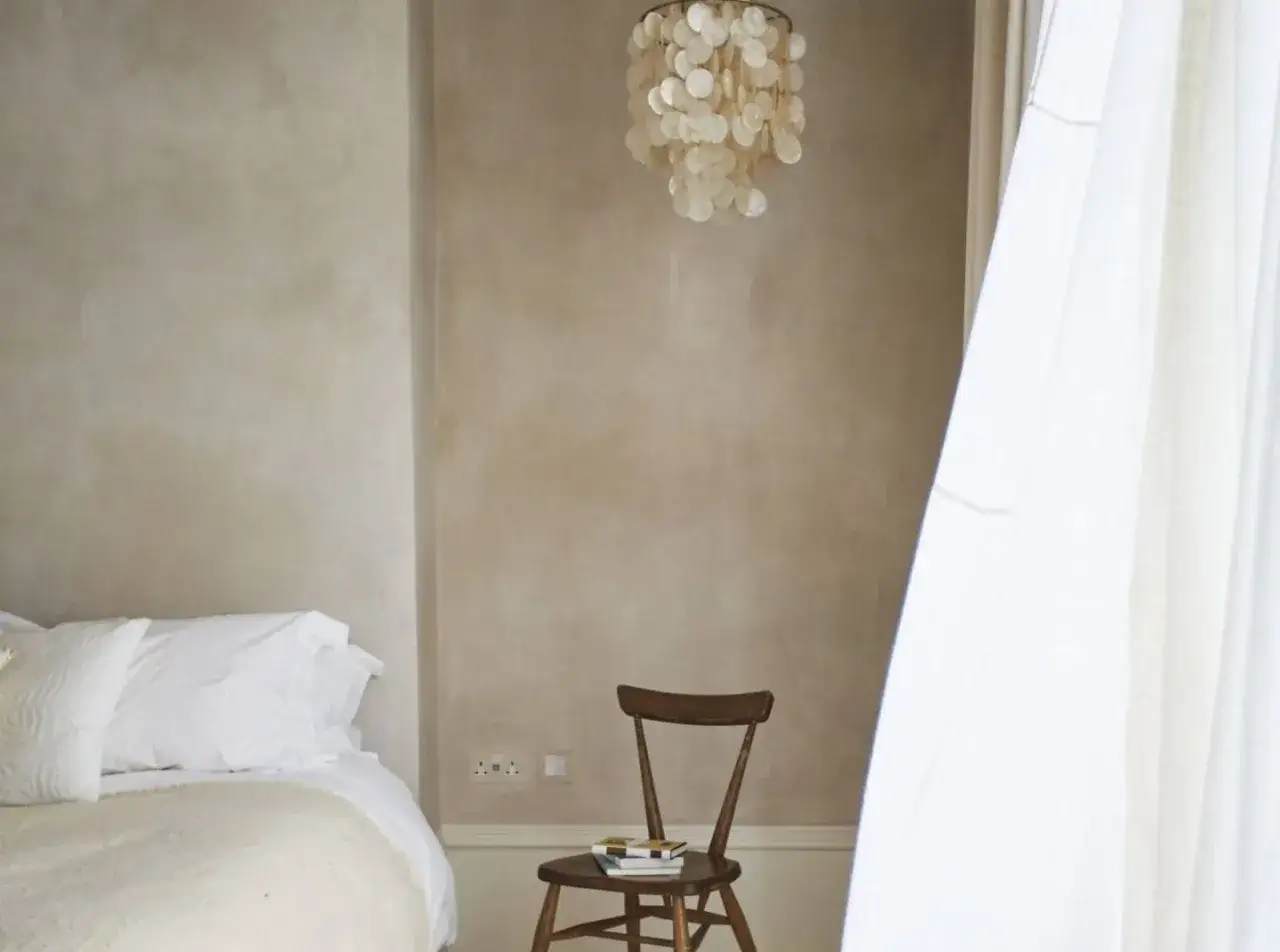 Parete con finitura pittura tamponato, letto bianco, sedia in legno con libri e lampadario di conchiglie.