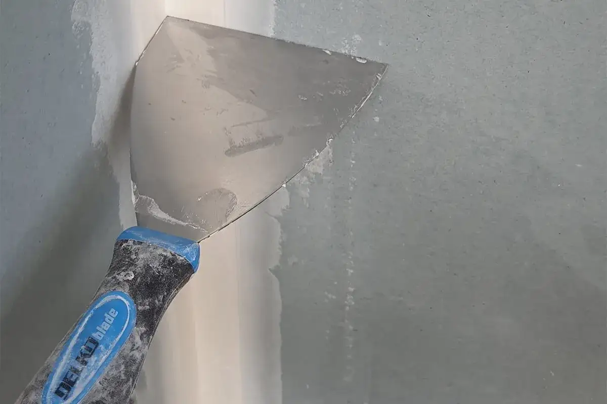 Una spatola metallica con manico blu applica lo stucco su un angolo di cartongesso, preparando la parete per la pittura.
