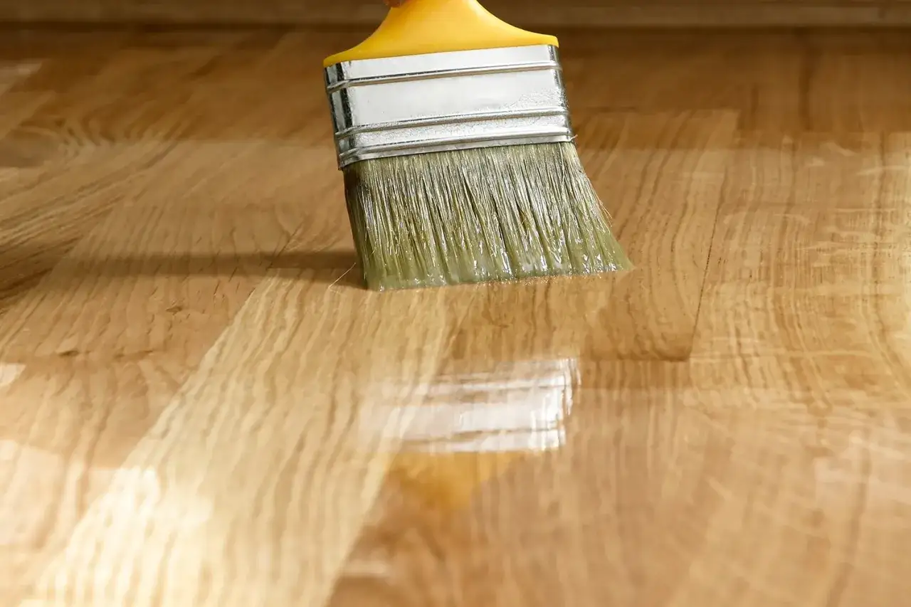 Pennello applica vernice trasparente su parquet in legno, creando un effetto lucido. L'obiettivo è opacizzare vernice lucida per un aspetto più naturale.