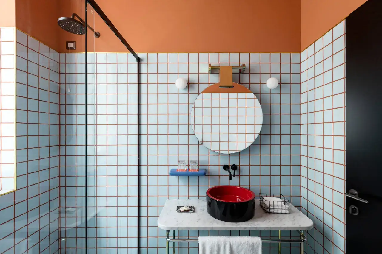 Bagno moderno con piastrelle azzurre e dettagli arancioni. Il lavabo nero e rosso, lo specchio rotondo e la doccia a vista suggeriscono una recente sostituzione piastrelle bagno.