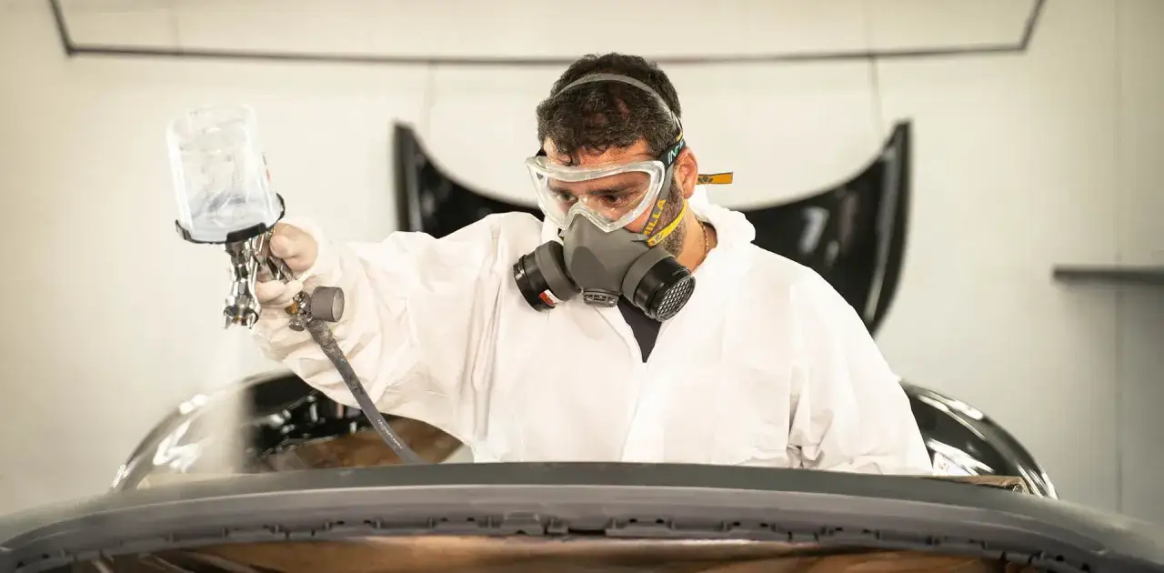 Operaio con tuta protettiva, maschera e occhiali spruzza vernici industriali su un pezzo di carrozzeria.