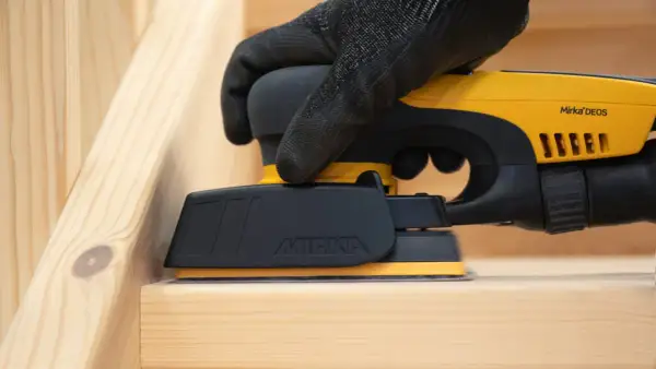 Mano guantata utilizza levigatrice elettrica per carteggiare legno grezzo, preparando la superficie per la finitura.