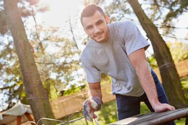 Uomo sorridente con guanti da giardinaggio lavora a un tavolo da giardino fai da te, immerso nel verde.