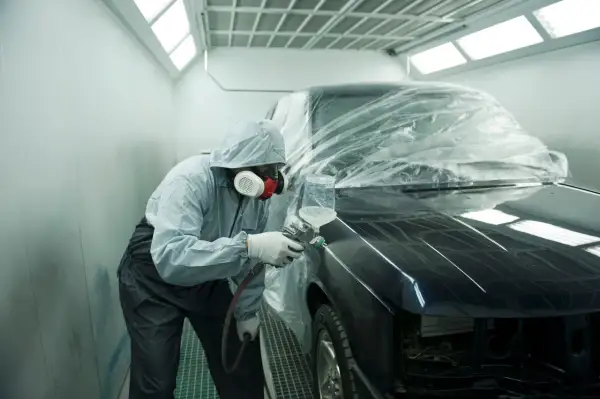 Un operaio in tuta protettiva e maschera spruzza vernice su un'auto scura, parzialmente coperta da un telo trasparente. L'auto sembra rovinata, in fase di restauro.