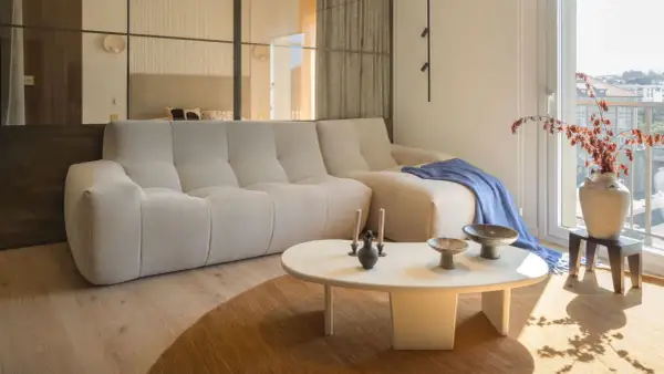 Divano chiaro e tavolino bianco su un tappeto di colore marrone. Sul tavolino, vasi e candele. Sul divano, una coperta blu.