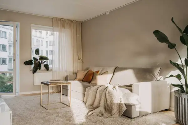 Un accogliente soggiorno con divano chiaro, cuscini color terra e una coperta morbida. Il tavolino dorato e la pianta verde aggiungono un tocco di stile. Pareti color marrone chiaro.