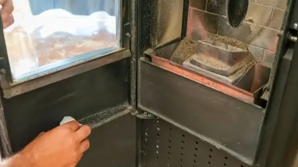 Mano che pulisce il vetro di una stufa a pellet, rimuovendo la cenere per una migliore visione del fuoco.
