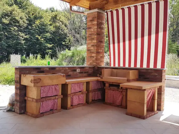 Cucina esterna in muratura fai da te con lavello, piano di lavoro e seduta. Tende a righe rosse e bianche proteggono lo spazio.