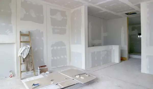 Interni in fase di finitura, con pareti in cartongesso pronte per essere stuccate. Una scala appoggiata al muro, pannelli e attrezzi a terra suggeriscono come fare un muro.